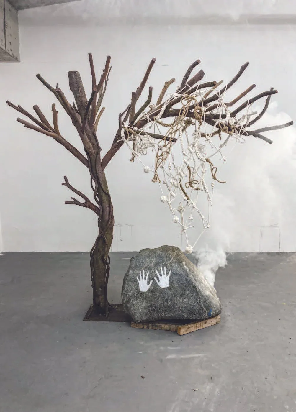 Installation of branch-built tree with sagging knotted rope net, a boulder marked by handprints, and drifting smoke — reflecting on the fragility of time and the necessity of living fully rather than merely recording.